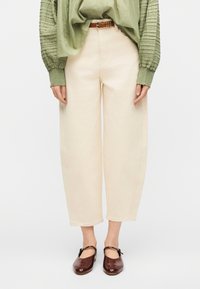 Γυναίκα που φοράει κρεμ παντελόνι με κανονική μέση, καφέ ζωνάρι, πράσινη μακρυμάνικη μπλούζα με υφή και σκούρα καφέ παπούτσια Mary Jane.