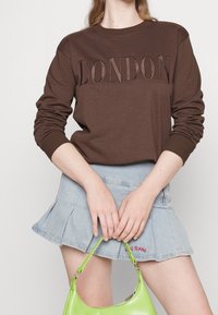 Brun sweatshirt med texten "LONDON" i upphöjd söm, kombinerad med en ljusblå jeanskjol och en klargrön handväska.
