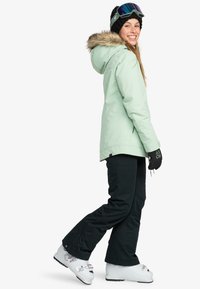 Mint green insulated parka with a fur-lined hood, paired with black ski pants and white ski boots, featuring mirrored goggles and black gloves.