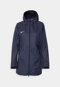 Giacca leggera blu navy con cappuccio. Presenta chiusura con zip, due tasche laterali e il logo Nike bianco sul lato sinistro del petto. Materiale resistente all'acqua.