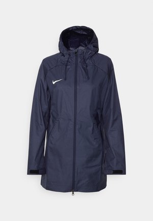Giacca leggera blu navy con cappuccio. Presenta chiusura con zip, due tasche laterali e il logo Nike bianco sul lato sinistro del petto. Materiale resistente all'acqua.