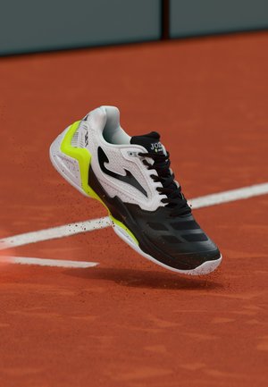 Black, white, and neon yellow sports shoe kicking up clay on an orange tennis court near a white line.