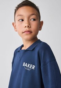 Marineblå polo skjorte med korte ærmer, med en krave og et hvidt logo tryk, der står "BAKER by Ted BAKER" på venstre bryst.