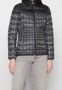 Svart pufferjacka med hög krage och framzipper, prydd med ett upprepande logomönster i vitt, med elastiska ärm- och en lätt textur.