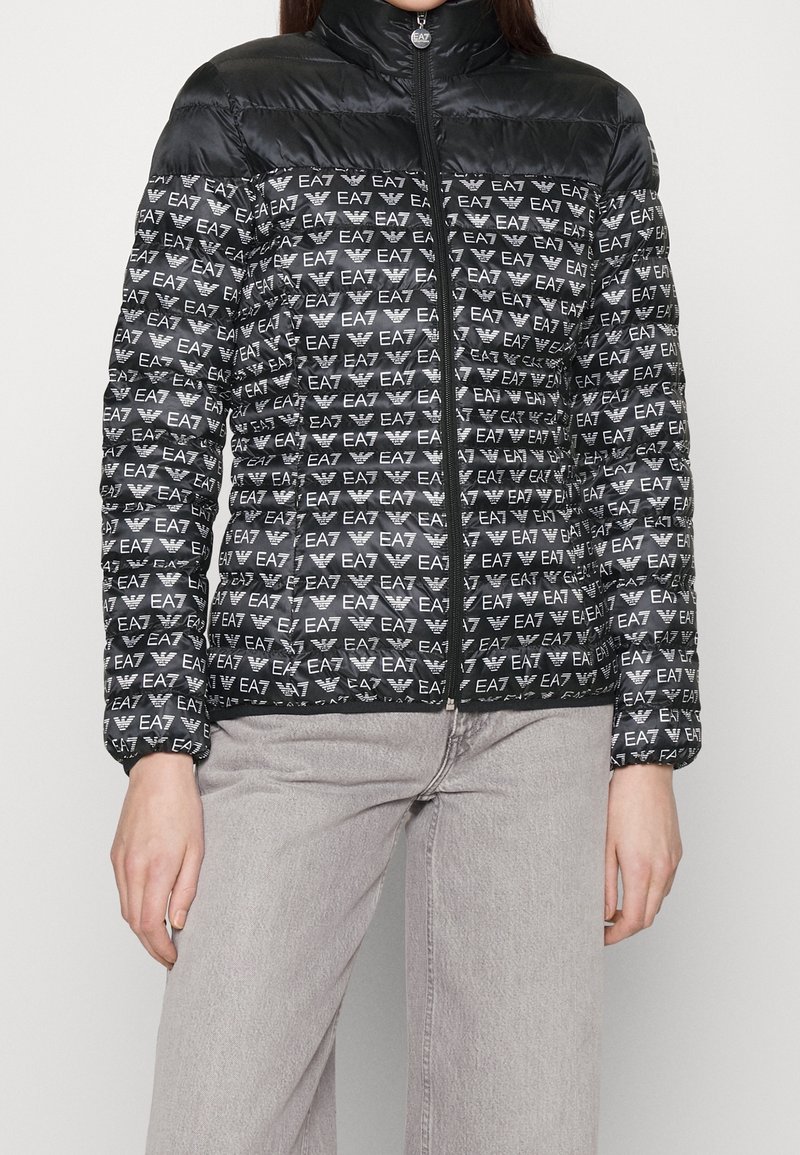 Svart pufferjacka med hög krage och framzipper, prydd med ett upprepande logomönster i vitt, med elastiska ärm- och en lätt textur.