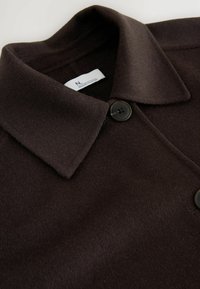 Manteau en laine marron foncé avec un large col et un grand bouton noir fixé près du haut.