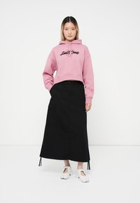 Giovane donna che indossa un hoodie corto rosa, una lunga gonna nera con tasche e scarpe bianche a punta divisa, in piedi contro uno sfondo bianco.