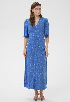 Femme debout portant une robe midi bleue à motifs floraux avec des manches longues jusqu'au coude et des sandales plates marron clair, regardant vers sa gauche.
