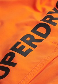 Tissu orange avec de grands caractères noirs écrivant "SUPERDRY". Texture lisse avec des coutures visibles le long du bord.