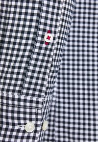Tela de camisa de cuadros en blanco y negro con puño abotonado, con botones blancos y detalles de costura en rojo. Textura suave, diseño ajustado.