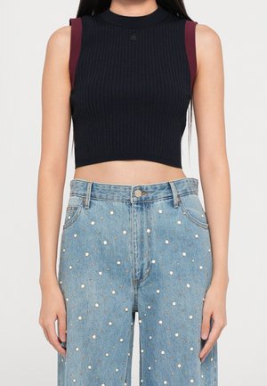 Vrouw draagt een mouwloze zwarte crop top met bordeauxrode schouderaccenten en een high-waisted blauwe spijkerbroek versierd met kleine parels en studs.