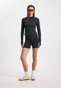 Musta urheilu takki, jossa on vetoketju kauluksessa, yhdistettynä samankaltaisiin pyöräilyshortseihin. Mallilla on päällään silmälasit ja vaaleat kengät, joissa on teksturoidut pohjat.