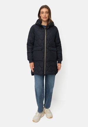 Woman standing wearing a black quilted hooded jacket, blue jeans, and white sneakers on a white background.