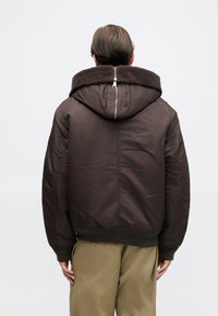 Καφέ μπουφάν με κουκούλα και επένδυση από γούνα, μπροστινό φερμουάρ, ριγέ μανσέτες και υφή, συνδυασμένο με ανοιχτό καμηλό παντελόνι.