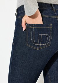 Jeans de mezclilla oscura con corte recto, que cuentan con un bolsillo trasero acentuado por un detalle de logo cosido en hilo beige. Tela texturizada visible.