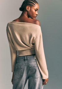 Beige off-the-shoulder sweater with a ribbed hem, paired with light gray high-waisted jeans featuring two back pockets and a faded texture.