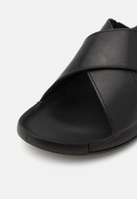 Sandalo in pelle nera con design incrociato, punta aperta e finitura liscia. La suola è piatta con un bordo leggermente strutturato.