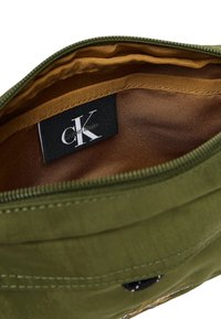 Interior de bolsa de tela verde oliva con forro marrón, con una etiqueta negra con el logo de Calvin Klein, textura suave y cierre de cremallera.