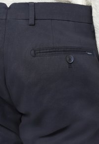 Calças azul-marinho feitas de tecido liso com um design ajustado, apresentando um bolso traseiro único com botão e etiqueta da marca.