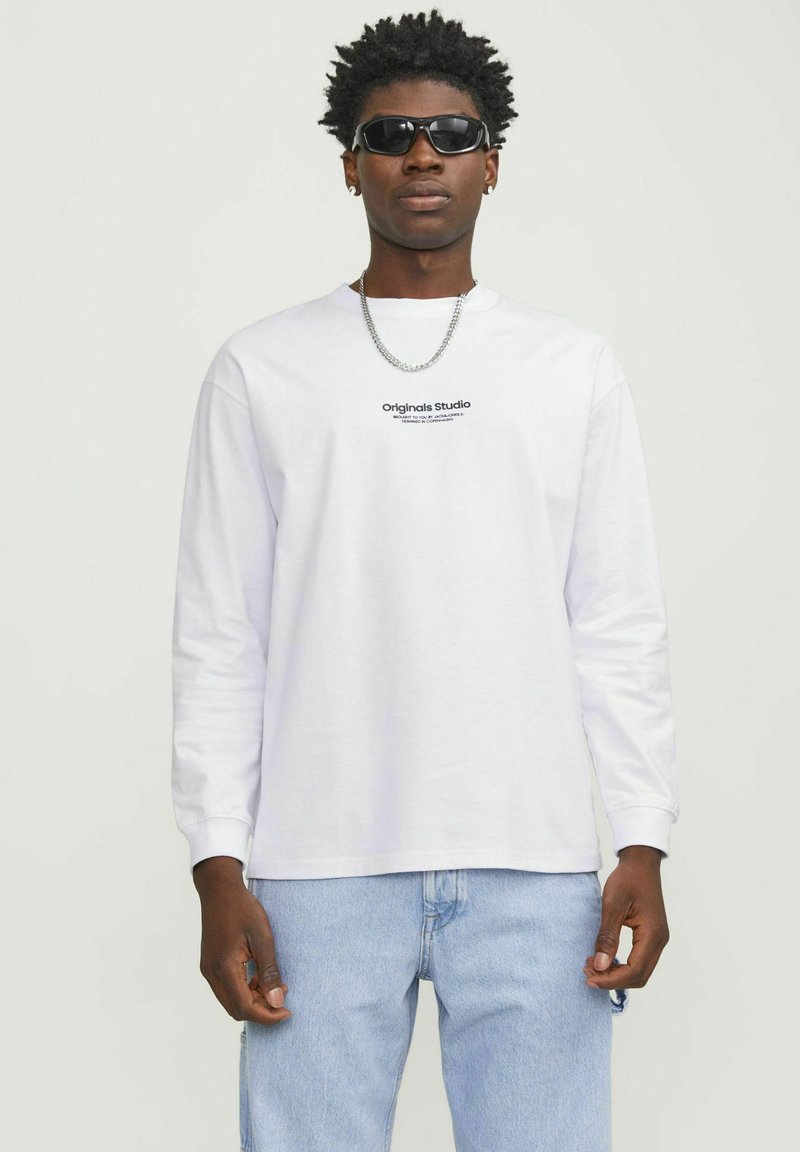 White long-sleeve t-shirt with a crew neckline and black text logo. Paired with light blue jeans and accessorized with sunglasses and a chain.