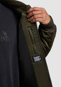 Olivegrüne Bomberjacke mit dunkel gefüttertem Innenleben. Verfügt über gerippte Bündchen, eine Reißverschlusstasche und ein im Innenfutter eingenähtes Markenlabel.