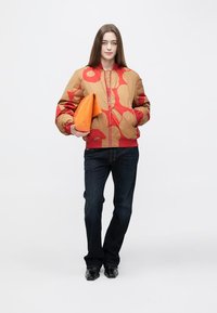 Obojestranska bomber jakna v bež barvi z rdečim cvetličnim vzorcem, kombinirana s temnimi jeans hlačami, v roki drži oranžno torbico, črni čevlji.