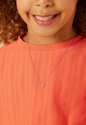 Zilveren ketting met een kleine bloemhanger, gedragen over een structuurtrui in koraal. Duidelijke details omvatten de schakels van de ketting en de vorm van de hanger.