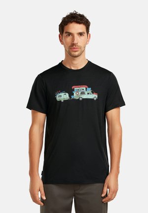 Mann trägt ein schwarzes T-Shirt mit einem Motiv eines Vintage-Camper-Trucks, der zwei Anhänger zieht und Outdoor-Ausrüstung auf dem Dach hat.
