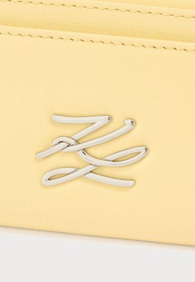Accessoire en cuir jaune à la texture lisse, avec un logo argenté proéminent au design élégant en cursive.