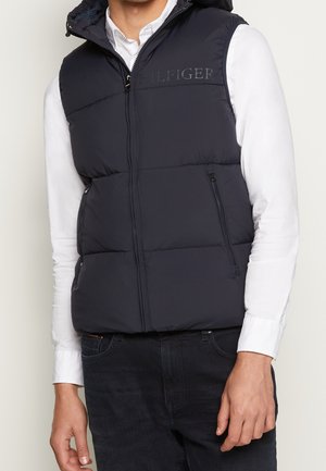 Mann trägt schwarze gesteppte ärmellose Weste über weißem Hemd mit Knöpfen und schwarzen Jeans, steht mit locker an den Seiten hängenden Armen.