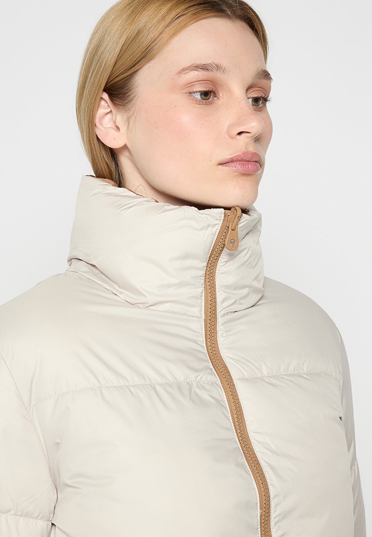 Chaqueta acolchada de color beige claro con un alto cuello, con una cremallera marrón y textura acolchada horizontal. Sin logotipos ni patrones visibles.