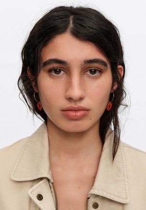 Joven mujer con cabello oscuro recogido, lleva una chaqueta beige y pendientes naranjas en forma de corazón, mirando hacia adelante con expresión neutral.