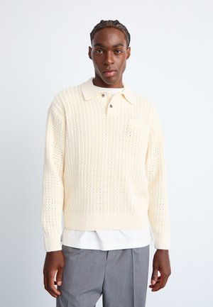 Cream-colored sweater with a textured knit design, featuring a collared neck and button accents. A front pocket is included.