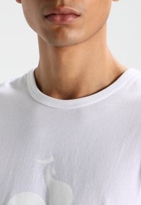 Lähellä otettu kuva miehen alahuulesta ja kaulasta, joka pitää päällään valkoista pyöreäkaulusta T-paitaa, jossa on hienovarainen vaalea kuosi.