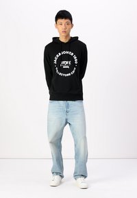 Schwarzer Kapuzenpullover mit einem weißen, runden Logo und Text, kombiniert mit hellblauen, weiten Jeans und weißen Sneakers. Das Modell steht vor einem schlichten Hintergrund.