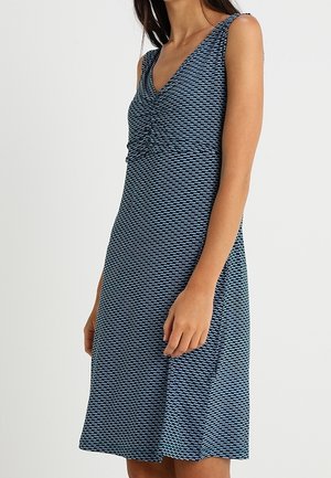 Frau trägt ärmelloses marineblaues Kleid mit hellblauem geometrischem Muster und V-Ausschnitt, steht vor einem schlicht hellen Hintergrund.