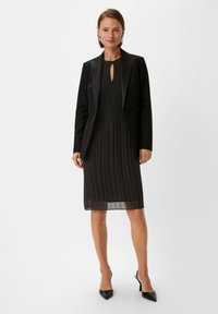 Schwarz plissee Kleid mit Schlüsselloch-Ausschnitt, getragen unter einem taillierten schwarzen Blazer, ergänzt durch spitze schwarze High Heels. Glattes Material und maßgeschneiderte Passform.