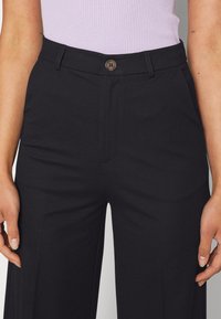 Czarne szerokie spodnie o gładkim wykończeniu, z zapięciem na guzik z przodu i paskami, umiejscowione nad jasnopurpurową bluzką.