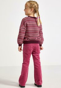 Strikket stribet sweater i bordeaux, pink og creme; parret med lyserøde fløjsbukser med vide ben. Modellen har fletninger og står sidelæns.