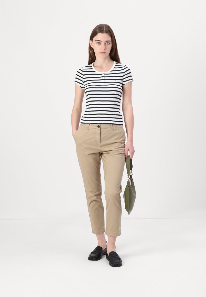 Gestreiftes schwarz-weißes Kurzarmshirt mit Henley-Ausschnitt, kombiniert mit beigen, taillierten Hosen und schwarzen Loafers, dazu eine grüne Tasche.