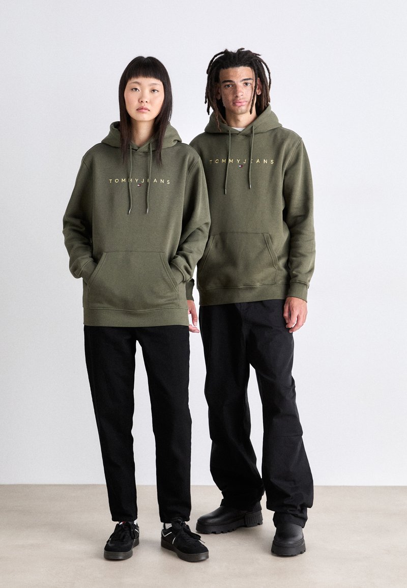Felpa verde con logo "TOMMY JEANS". Modello in coppia, uno in pantaloni neri, l'altro in pantaloni neri a vestibilità rilassata. Entrambi indossano calzature nere.
