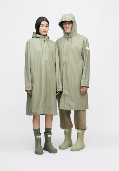 Kaks isikut kannavad kergest, oliivrohelisest kangast vihmamantleid, millel on kapuuts, koos sobivate vihmavate jalatsitega, millel on logo ja tekstuurivalikud.