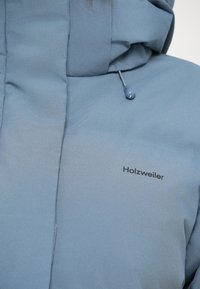 Svijetloplava izolirana jakna s visokim ovratnikom, kapuljačom s podesivim špagom i crnim logom "Holzweiler" sprijeda. Glatka, mekana tekstura.