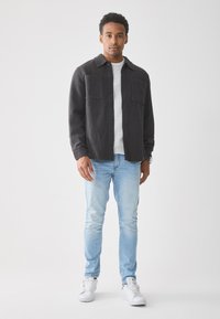 Camisa de franela gris oscuro con diseño de botones, dos bolsillos en el pecho, que se lleva sobre una camiseta gris claro. Jeans azul claro y zapatillas blancas completan el look.
