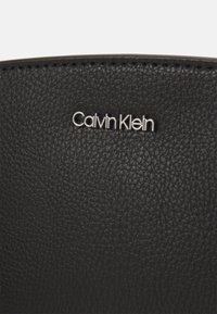 Schwarzes strukturiertes Leder mit einem silbernen "Calvin Klein"-Logo. Die Oberfläche weist ein genarbtes Muster und präzise Nähte entlang der Kanten auf.