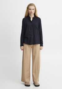 Camicia button-up blu navy con taschino frontale, abbinata a pantaloni beige largi e calzature grigie. Tessuto morbido con vestibilità rilassata.