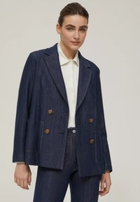 Giubbotto doppiopetto in denim scuro con bottoni marroni, due tasche frontali, rever a punte e una vestibilità comoda, indossato sopra una camicia bianca.