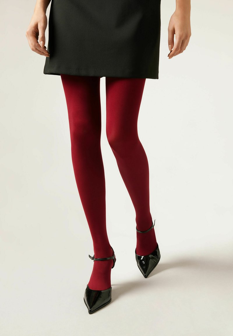 Burgunderfarbene Strumpfhosen kombiniert mit einem schwarzen Kleid und schwarzen Lackleder-Pumps mit Knöchelriemen. Glatte Textur, figurbetontes Design.
