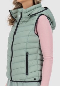 Gilet imbottito verde chiaro con cappuccio, caratterizzato da cuciture orizzontali, zip nera e tasche laterali. Indossato sopra una maglia rosa a maniche lunghe.