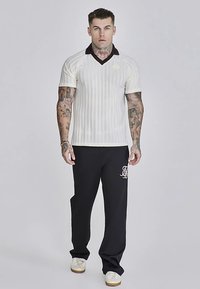 Kremno in rjavo črtasta polo majica s kratkimi rokavi in V-izrezom, kombinirana s črnimi trenirkami z belim logotipom. Bele superge.